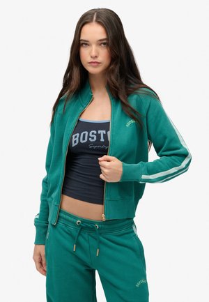 Veste zippée bleu sarcelle avec des poignets et un col côtelé, ornée d'accents dorés et de rayures latérales. Portée sur un débardeur bleu marine comportant l'inscription "BOSTON".