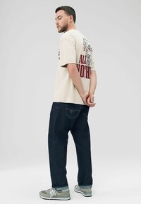 T-shirt beige à manches courtes avec un design graphique rouge au dos, associé à un jean bleu foncé et des chaussures de sport grises, se tenant avec les mains derrière le dos.