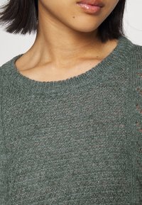 Grön stickad tröja med lös väv, rund halsringning och strukturerat mönster. Har breda, öppna stygn för andningsförmåga.