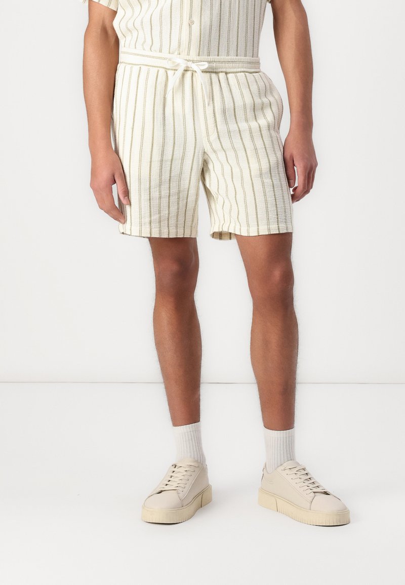 Shorts pour hommes en tissu ivoire texturé avec des rayures verticales vertes, dotés d'une taille élastique et d'un cordon de serrage. Associés à des baskets beiges.