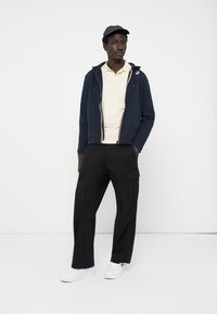 Marineblå zip-up hættetrøje med kontrastlynlås, lys gul polo t-shirt, sorte cargo bukser og hvide sneakers; modellen har en sort kasket på.