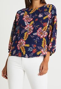 Blouse florale marine avec un motif vif de fleurs roses, jaunes et vertes. Coupe ample avec des manches trois quarts et tissu doux.