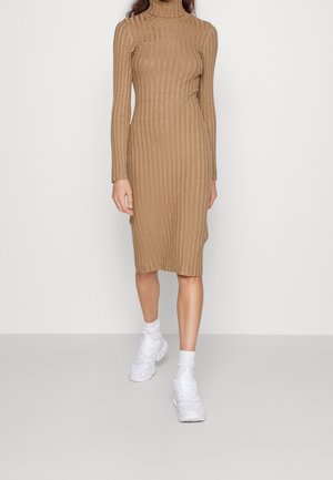 Vrouw draagt een aansluitende, geribde, camelgekleurde coltrui midi-jurk, gecombineerd met witte sneakers en sokken, staand tegen een effen achtergrond.