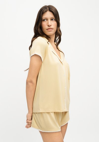 Femme aux longs cheveux bruns porte une chemise beige à manches courtes boutonnée et un short assorti avec un liseré blanc, debout les mains derrière le dos.