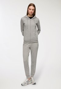 Felpa grigia con zip, dotata di lacci e due tasche frontali, abbinata a pantaloni della tuta grigi coordinati. Il modello indossa scarpe da ginnastica argento.