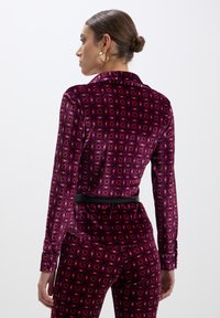 Completo di giacca e pantaloni in velluto di un rosa intenso con motivi geometrici. Presenta un colletto, maniche lunghe e un cinturino nero in evidenza in vita.