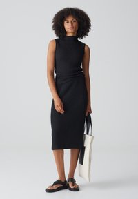 Zwarte mouwloze jurk van textuurstof, knielang ontwerp en hoge halslijn. Model houdt een crèmekleurige tote bag vast met zwarte accenten.