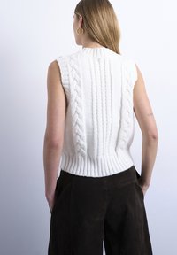 Femme aux cheveux blonds raides portant des boucles d'oreilles créoles en or, un pull sans manches en coton torsadé blanc et un pantalon marron foncé, les mains dans les poches.