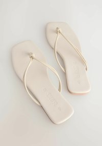 Ljust beige platta sandaler med fyrkantig tå och tunna gummisnoddar knutna i mitten. Sulan är slät med subtilt varumärke.