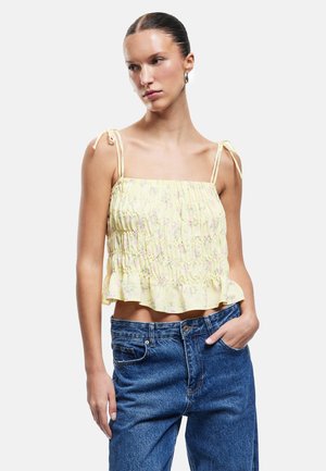 Kvinde iført en gul blomsterprintet smock-crop top med bindebånds stropper og højtaljede blå jeans, står med den ene hånd i lommen.