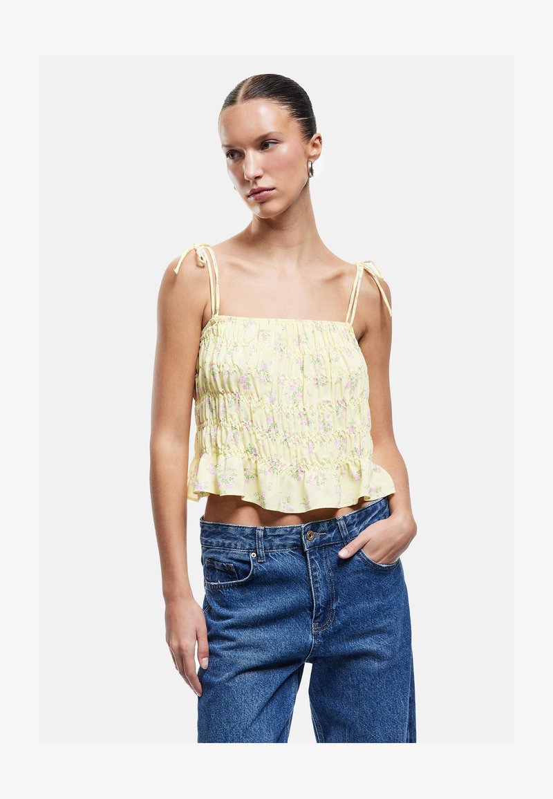 Femme portant un crop top smocké jaune à fleurs avec des bretelles à nouer et un jean bleu taille haute, debout avec une main dans la poche.