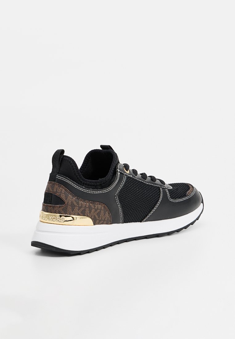 Zapatilla de lona baja en negro y marrón con suela blanca, paneles de malla, cordones negros y un detalle en dorado en el talón con el nombre de la marca.