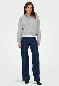Femme portant un sweat à capuche court gris clair, un jean droit bleu foncé et des bottes à talons aiguilles noires, se tenant devant un fond uni.