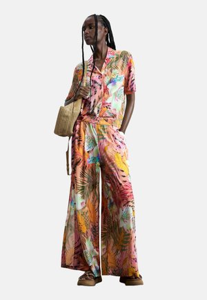 Donna che indossa una camicetta floreale colorata a maniche corte e pantaloni a gamba larga, tiene in mano una borsa di paglia intrecciata, in piedi davanti a uno sfondo neutro.