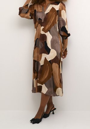 Vrouw draagt een midi-jurk met een abstract patroon in bruin, beige en zwart, met opgeblazen lange mouwen en zwarte slingbacks met hak.