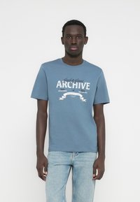 Plava pamučna majica kratkih rukava s okruglim izrezom, s bijelim ispisom "ARCHIVE" i dekorativnim detaljima sprijeda.