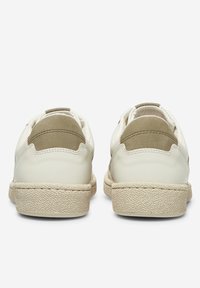 Pár krémových tenisek zobrazených ze zadu, s texturovanými béžovými podrážkami a olivově zelenými suede patními prvky.