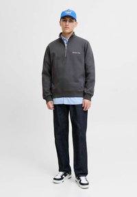 Sudadera gris de media cremallera con logotipo bordado, superpuesta a una camisa a rayas azul claro, combinada con jeans oscuros y zapatillas negras y blancas.