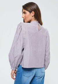 Femme de profil portant un chemisier rayé vertical violet et blanc avec des manches bouffantes et un jean bleu.