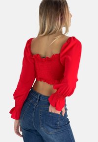 Top corto rojo con mangas abombadas, cuerpo fruncido y bordes con volantes. Combinado con jeans azul de tiro alto, destacando una silueta ajustada.