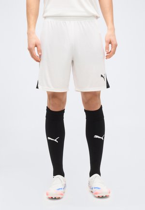 Apatinė kūno dalis dėvint balčius Puma sportinius šortus, juodas aukštas Puma kojines iki kelių ir baltus futbolo batelius su rožiniais ir mėlynais akcentais.