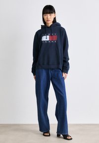 Donna che indossa un hoodie blu navy con il logo Tommy Jeans, jeans azzurri larghi e sandali con tacco aperti neri, in piedi contro uno sfondo chiaro e neutro.