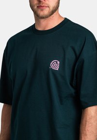 Camiseta de algodón en verde azulado oscuro con un logo en espiral rosa en el lado izquierdo del pecho. Mangas cortas y cuello redondo, con una textura suave.
