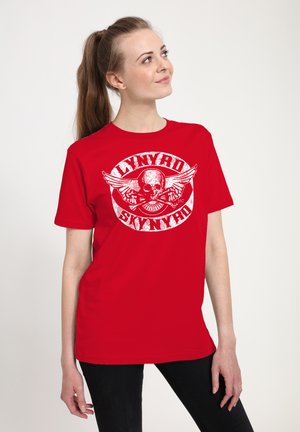 Ung kvinde med hestehale iført en rød Lynyrd Skynyrd T-shirt, kigger opad og står mod en ensfarvet lys baggrund.