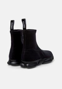 Botas negras hasta el tobillo hechas de tela elástica con un diseño tipo calcetín. Presentan una suela gruesa y texturizada, así como lengüetas en el talón con la marca.