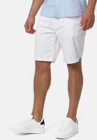 Pantaloni corti bianchi in cotone con orli arrotolati, abbinati a sneakers bianche con un accento nero. La camicia ha strisce verticali blu e bianche.