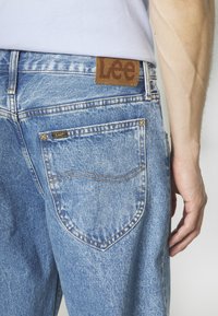 Světle modré džíny z denimu s hnědou koženou nášivkou na zadní straně, které mají dvě kapsy s žlutým prošíváním a měkkou texturu.