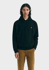 Čierna mikina s kapucňou z mäkkej látky s predným vreckom, nastaviteľnou šnúrkou na kapucni a malým oranžovým logom na hrudi.