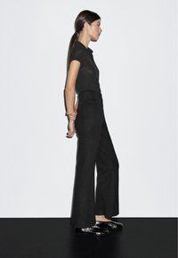 Zwarte poloshirt in combinatie met wijdvallende zwarte broek en glanzende zwarte flats, gepresenteerd tegen een plain witte achtergrond.