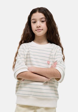 Jeune fille aux longs cheveux ondulés portant un pull rayé blanc et bleu clair, les bras croisés devant un fond uni.