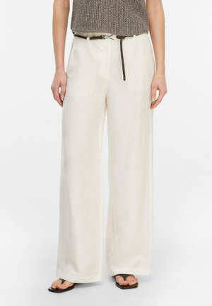 Dorosły nosi białe spodnie z wysokim stanem i szerokimi nogawkami, brązowy pleciony pasek, brązowy bezrękawnik dzianinowy oraz czarne sandały z kwadratowym noskiem.