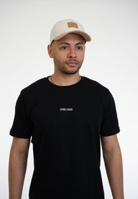 Fekete póló "JOHNNY URBAN" felirattal fehér színben, bézs sapkával párosítva, amelynek elején egy texturált bőrpötty található.