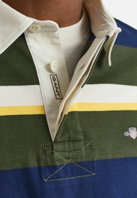 Camisa polo de manga corta con cuello en verde, azul marino y blanco con rayas horizontales, que presenta un logotipo de GANT y botones en el escote.