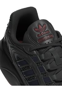 Scarpe sportive nere con una tomaia in mesh testurizzato, riflessi lucidi e un logo Adidas ben visibile sulla linguetta. Lacci e dettagli di design di supporto in evidenza.