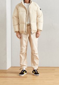 Beige Daunenjacke mit hohem Kragen, schwarzen Akzenten, cremefarbenem Hemd, beigen, schmal zulaufenden Hosen und schwarzen Turnschuhen mit gelben Streifen.