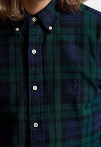 Vihreä ja tummansininen ruudullinen nappikauluspaita, jossa on brodeerattu logo rinnassa, käytössä olalla vaaleahius henkilö.