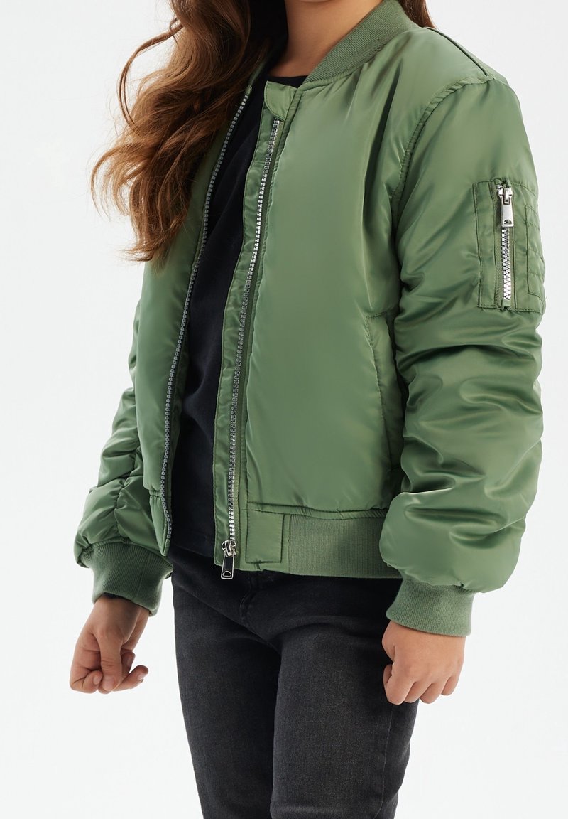 Fille aux longs cheveux portant une veste bomber verte par-dessus un t-shirt noir et un jean foncé, sur un fond clair uni.