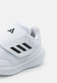 Bílá sportovní obuv s texturovaným svrchním materiálem, páskem na suchý zip a černými doplňky. Má zaoblenou špičku a pružnou gumovou podrážku.