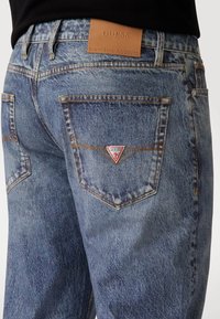 Vaqueros de denim azul claro con un parche de cuero con el logotipo de GUESS, logotipo en triángulo en el bolsillo trasero y un diseño clásico de cinco bolsillos con detalles de costura.