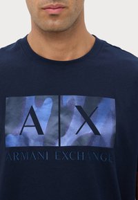 T-shirt in cotone blu navy con stampa grafica con "AX" in lettere scure e "ARMANI EXCHANGE" in testo ricamato nella parte inferiore.
