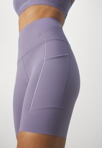 Gros plan d’une personne portant un short de sport lavande taille haute avec poches latérales et un soutien-gorge de sport assorti.