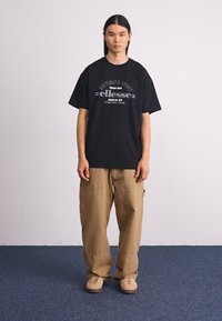 Svart oversized t-skjorte med "ellesse" logo i hvitt og grått, kombinert med sandfargede baggy cargo-bukser og beige joggesko på et blått teppe.