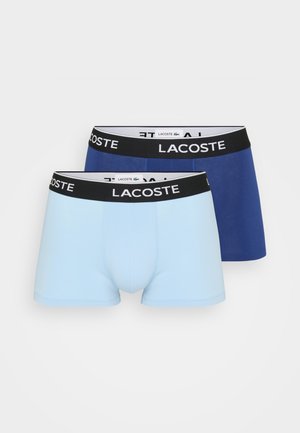 Dois pares de boxers Lacoste: um azul claro e um azul-marinho, apresentando uma cinta preta com o nome da marca em letras brancas.