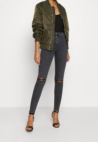 Olivgrön bomberjacka med silverdragkedja, kombinerad med höga svarta tajta jeans med knärivor och svarta klackskor med rem runt ankeln.