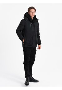 Uomo che indossa un parka isolato nero con cappuccio, pantaloni cargo neri e stivali neri con lacci, in piedi davanti a uno sfondo bianco uniforme.