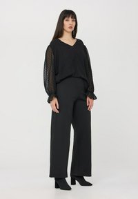 Blusa nera trasparente con pois testurizzati, scollatura a V e maniche a sbuffo; abbinata a pantaloni neri a vita alta e stivaletti con tacco a blocco.